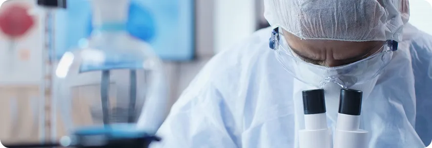 Laboratory technician in a protective gown, cap and goggles looking through a microscope